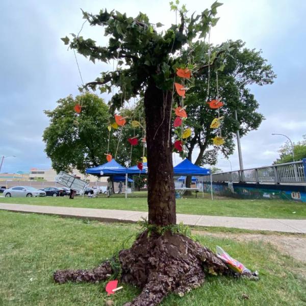 A tree in the middle of an open community space, with small pieces of red, orange and yellow construction paper tied with string to hand down from its branches.