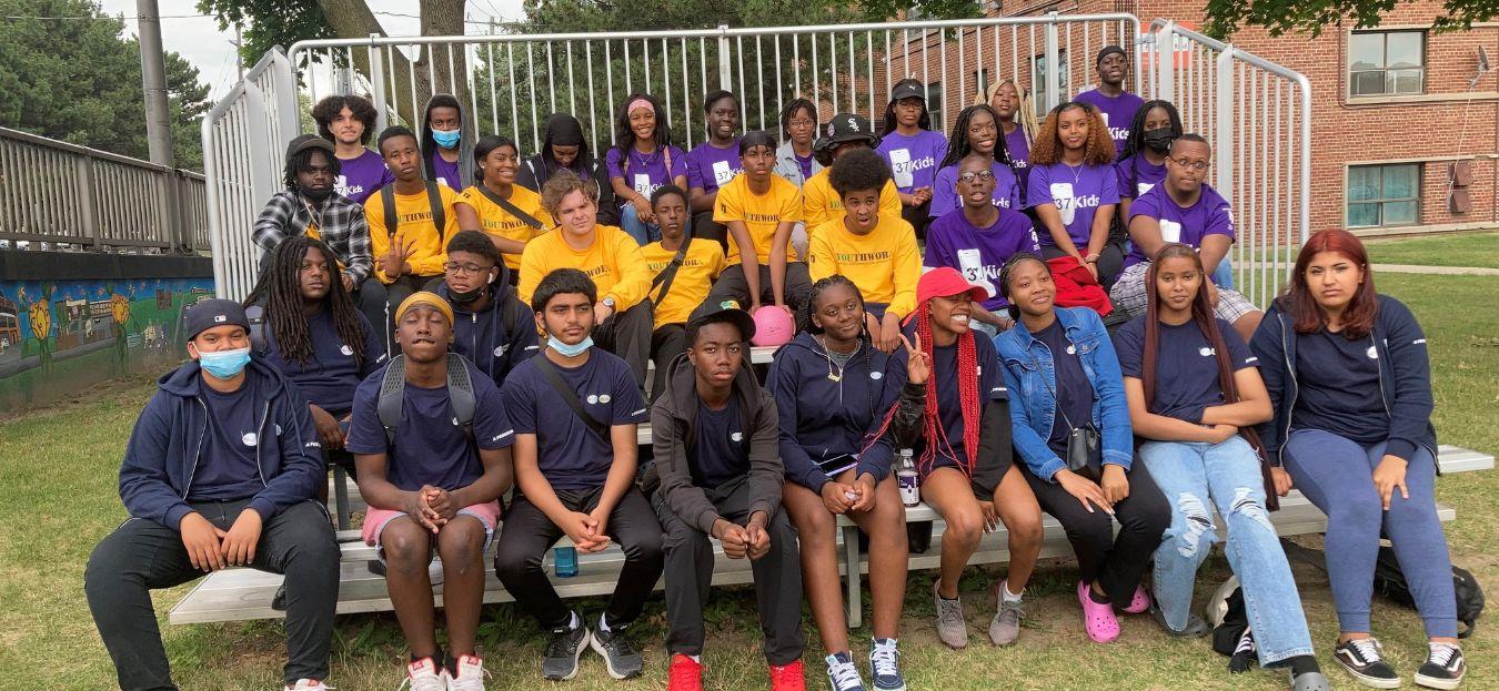 A group of participants from the YouthWorx( youth in yellow shirts) 37 Kids ( youth in purple shirts) and Revite Nerd (youth in dark blue) sitting on benches posing for the camera.