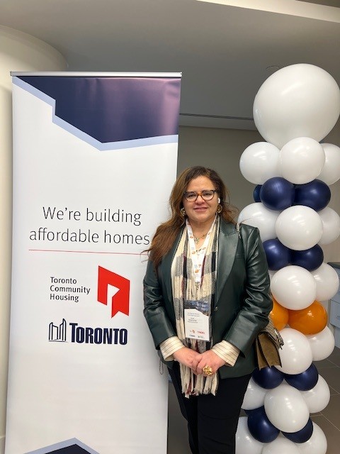 Woman with glasses, green jacket and a scarf on, standing besides banner and balloons
