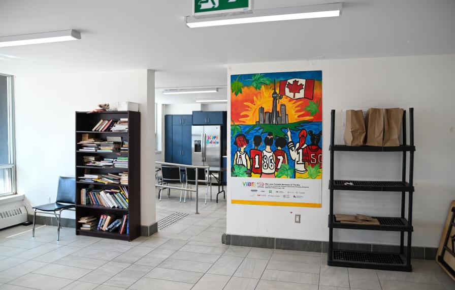 Community common room with a shelf full of books, colourful painting on the wall, leading into a kitchen space