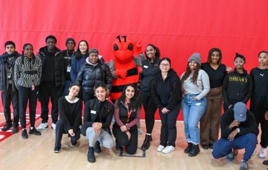 A group of students stand in front of a red wall. There is a red bee mascot in the middle.