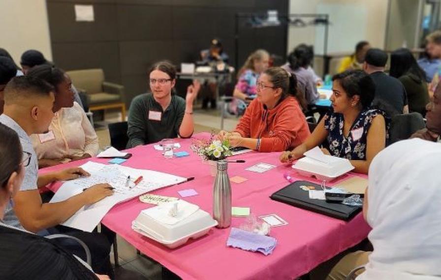 Diverse group of people at a table. Pink table cloth and papers line the table. 