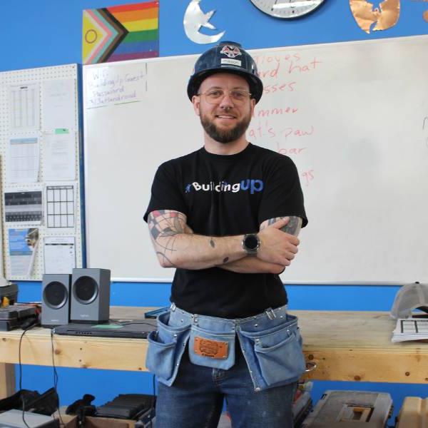 Person posing with arms crossed wearing tool belt and hard hat in the Building Up workshop.