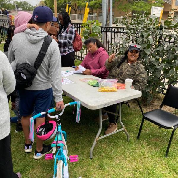 Two women sitting at a table outside, checking a list and giving out tickets, while other community residents line up.