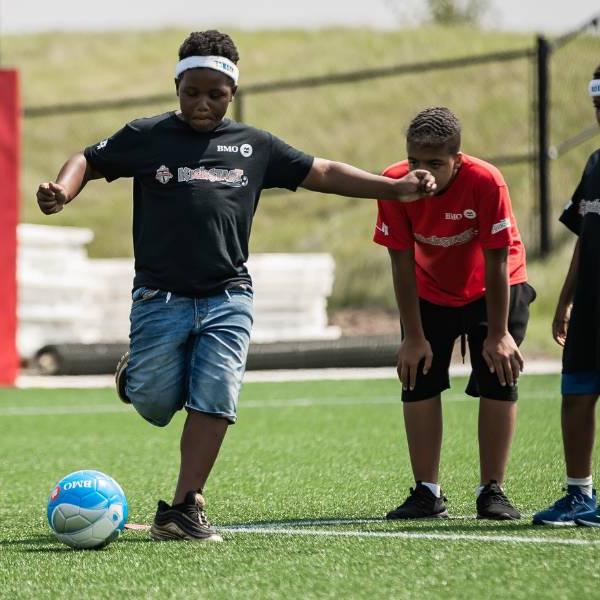 A boy kicking a soccer ball with two other participants waiting to take their turn.