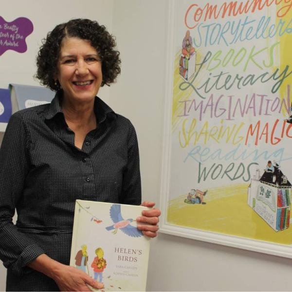 Mary Ladky holding the book 'Helen's Birds' and standing beside a poster with the words community, storytellers, books, literacy, imagination, sharing, magic, reading, words.