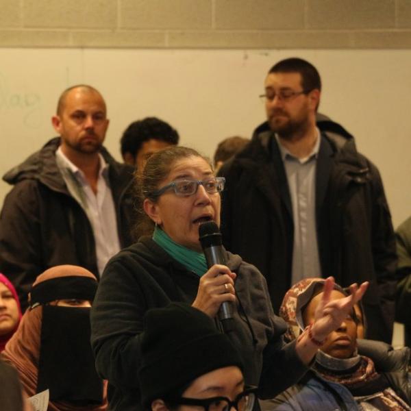 A woman wearing a black sweater and green scarf asking questions at the community meeting. 