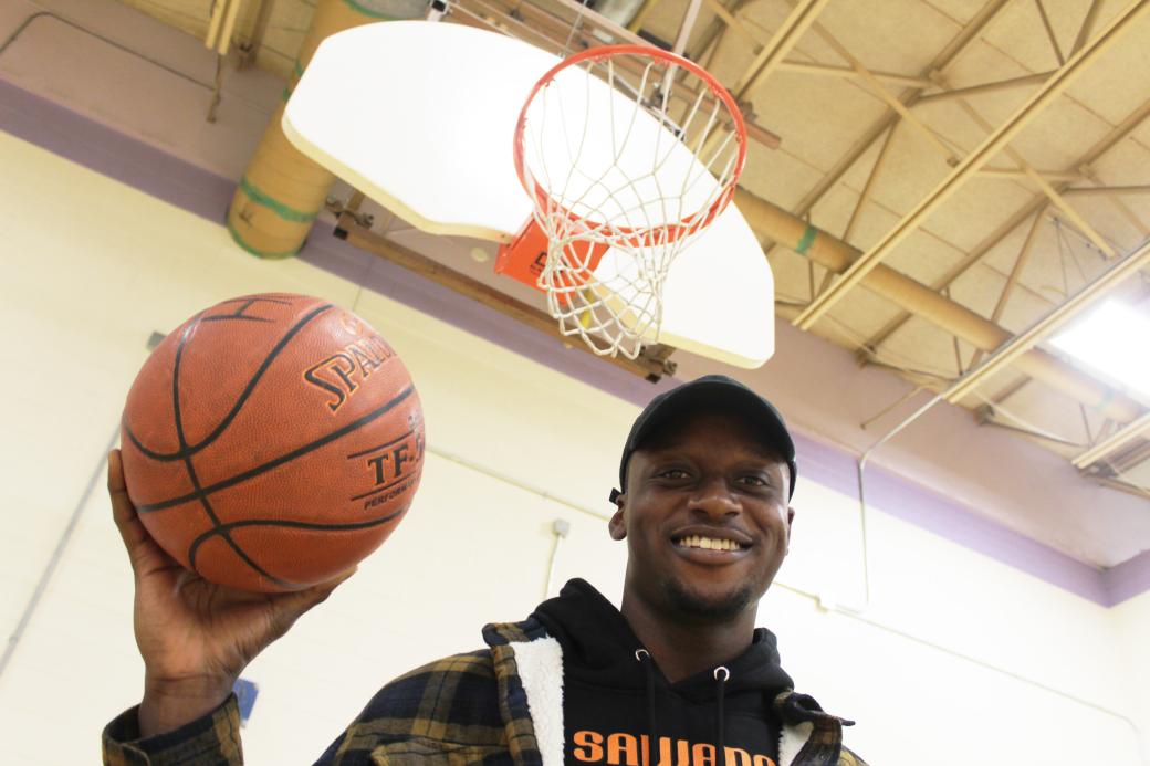 Man standing under a basketball net, holding a basketball in one hand, looking down at the camera and smiling