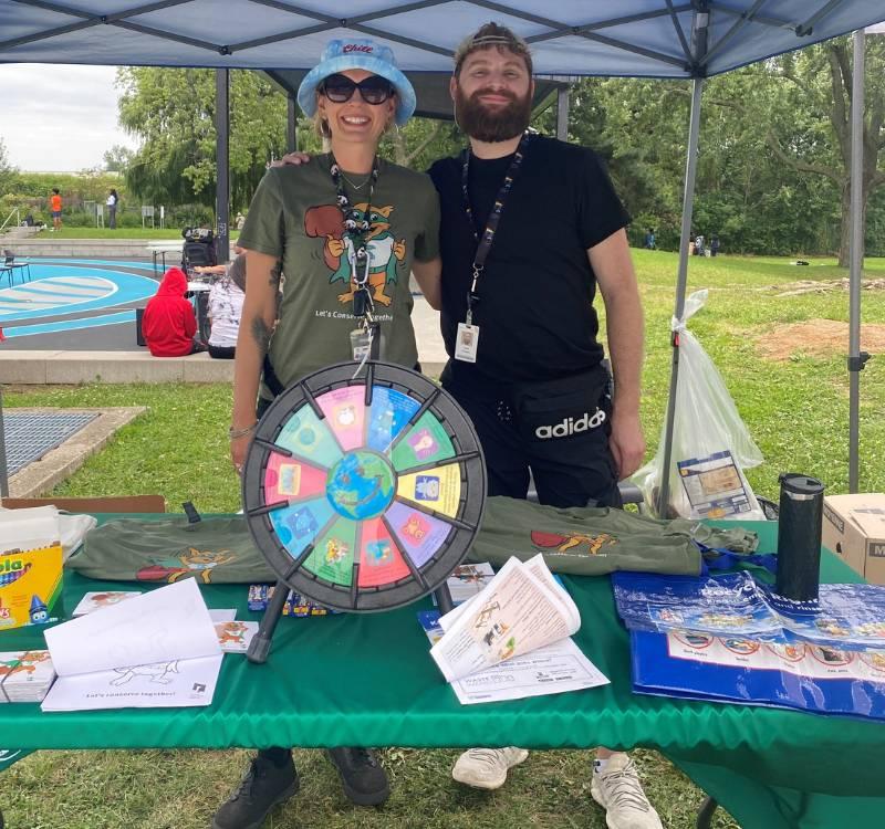 Two TCHC Conservation team staff stand behind a table at a community event. The table contains a prize wheel, coloring sheets and more promo items to help people learn about the importance of conservation.