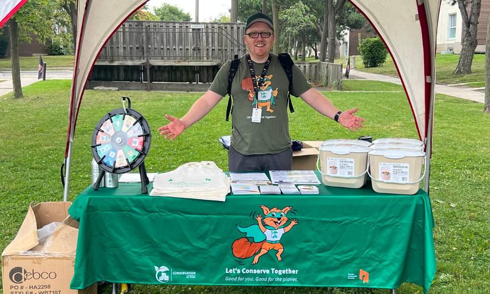 A member of the TCHC Conservation team stands behind a table under a tent in an outdoor greenspace. The table contains activities, promotional items and a prize wheel.