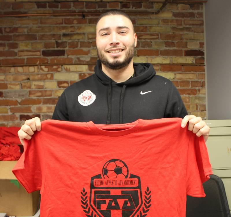 A man holding a red t-shirt with a logo on front featuring a shield and a soccer ball.