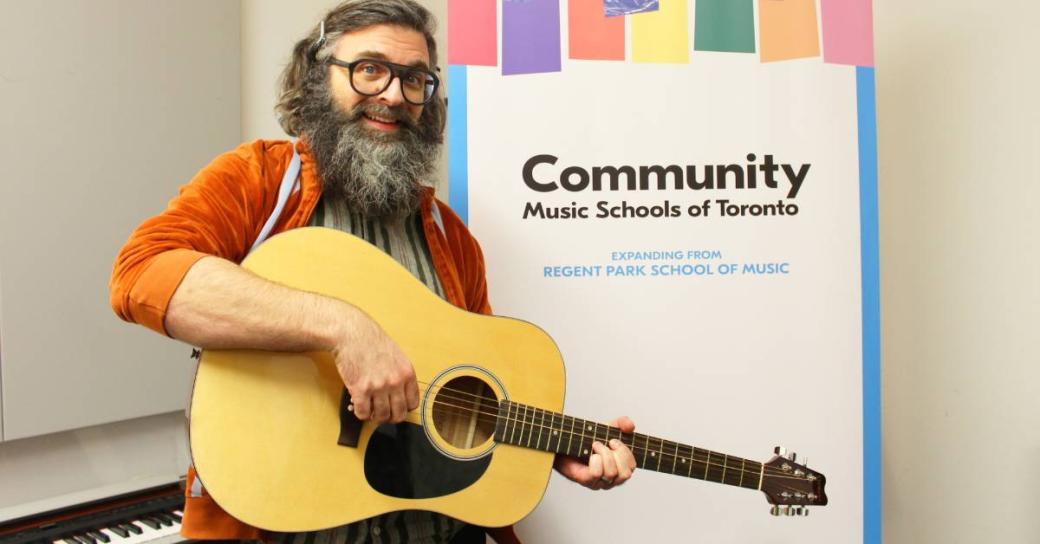 A man holding a guitar standing in front of a banner which says "Community Music Schools of Toronto".