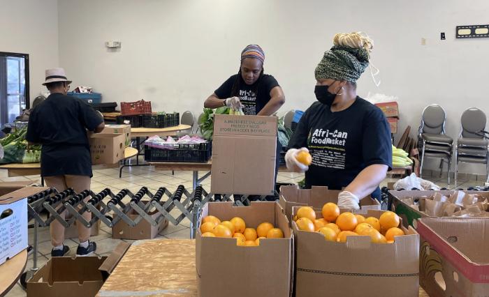 Three women in black shirts are seen stuffing boxes full of oranges.
